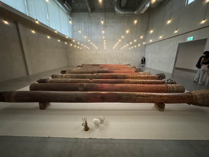 A row of old wrapped temple columns laid out on a pale floor in a bare concrete gallery hall at dib Bangkok, small offering vessels placed before them.