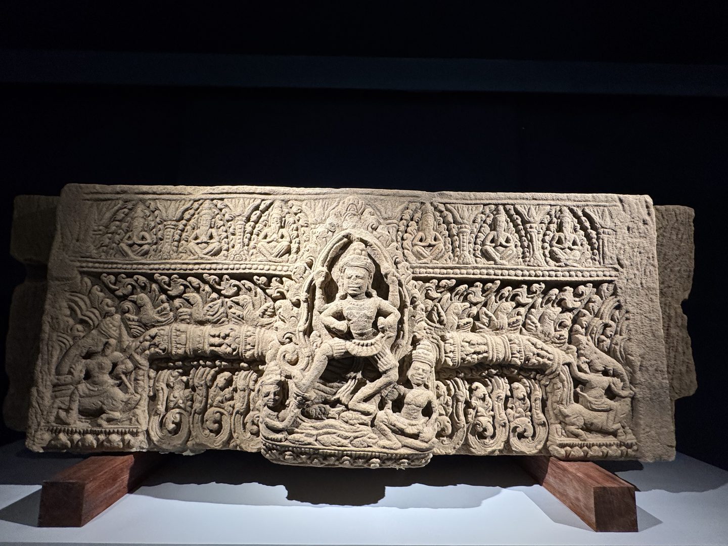 A carved Khmer sandstone lintel on display at the Phimai National Museum, a central deity figure framed by intricate foliate carving